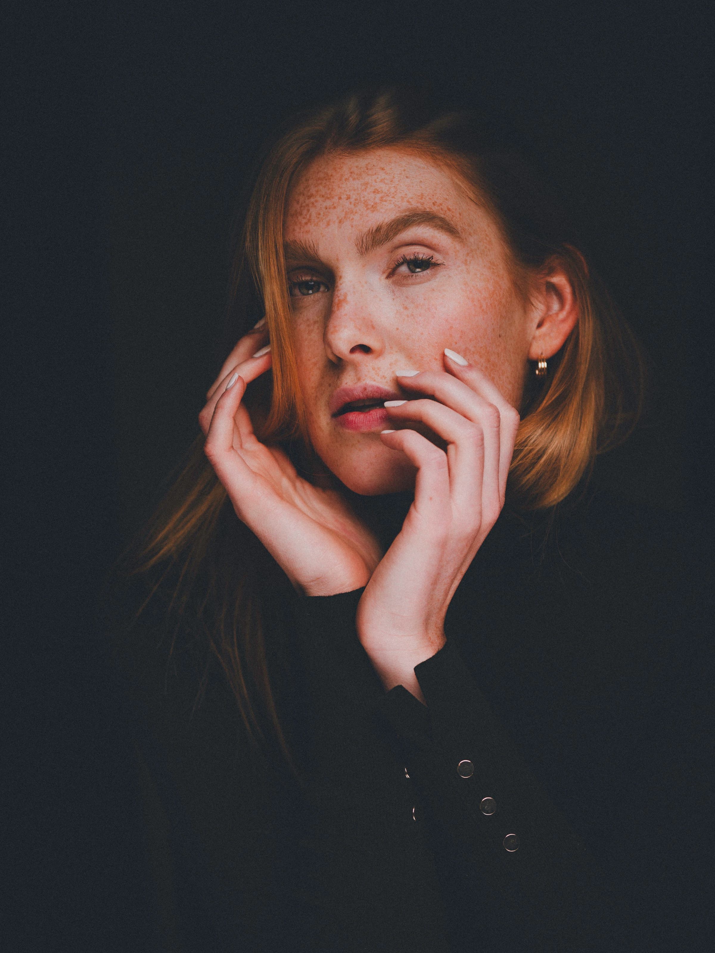 Intimate portrait of Fabia gently holding her face with both hands, looking directly at the viewer. Her freckles are highlighted by soft dramatic lighting against a deep black backdrop.